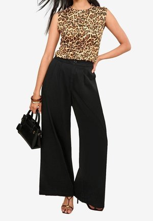 Donna che indossa una canotta senza maniche con stampa leopardata, pantaloni neri a vita alta e gamba larga, borsa nera e sandali marroni con cinturini.