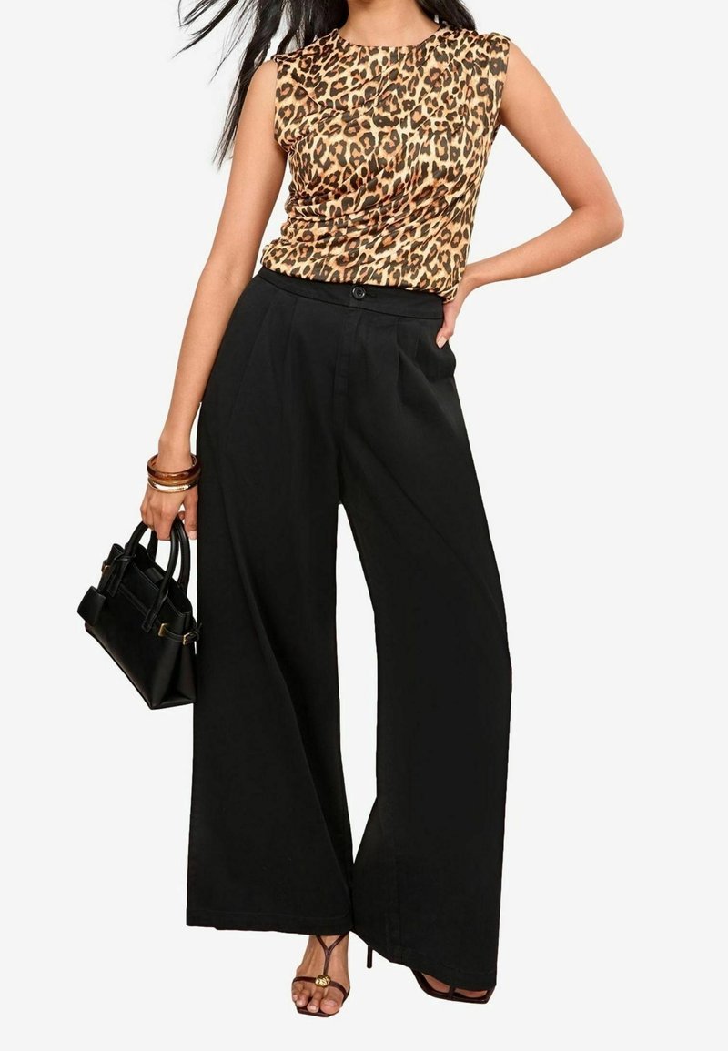 Femme portant un top sans manches imprimé léopard, un pantalon noir taille haute à jambes larges, un sac à main noir et des talons marron à brides.
