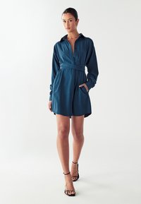 Turkizna obleka s kraticami in gumbi spredaj, dolgimi rokavi in pasom za zavezovanje. Ima dve stranski žepi in gladko teksturo.