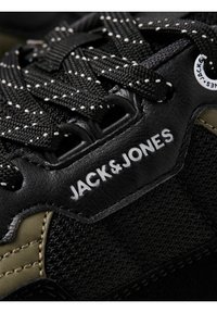 Gros plan d'une basket noire avec un tissu texturé, des lacets noirs et blancs à pois, et "JACK & JONES" imprimé en blanc sur un détail en cuir.