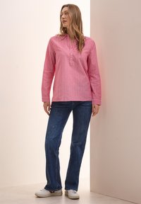 Camicia a maniche lunghe a righe rosa e bianche con scollo a V e dettagli con bottoni, abbinata a jeans svasati blu scuro e sneakers bianche.
