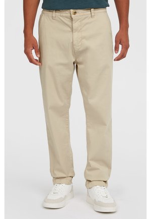 Mężczyzna ubrany w beżowe chino i białe trampki, stojący z ramionami swobodnie opuszczonymi wzdłuż ciała na jednolitym tle.