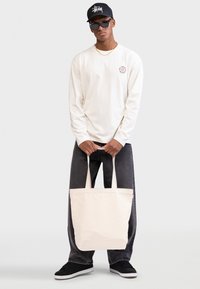 Giovane uomo che indossa un cappellino nero e occhiali da sole, maglia a maniche lunghe color crema, jeans neri, che tiene una grande borsa a tracolla beige davanti a uno sfondo bianco.