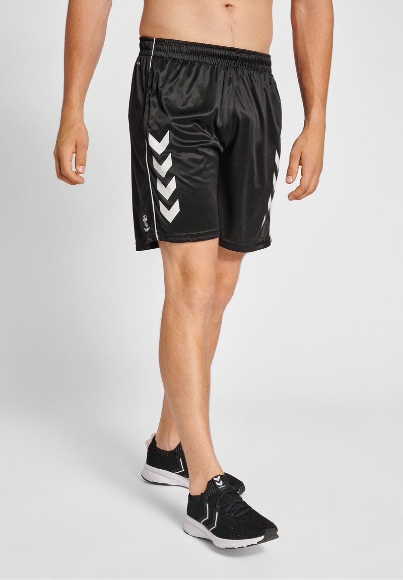 Homme portant un short de sport noir avec des motifs en chevron blancs et des chaussures de course noires avec des semelles blanches, debout sur un fond uni.