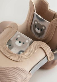 Încălțăminte cu exterior moale din cauciuc bej, cu accente gri și două panouri textile cu imprimeuri cu urși. Textură netedă cu bretele flexibile.