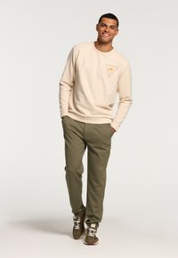 Beige sweatshirt with triangular graphic in yellow and orange, paired with green joggers and brown sneakers, standing on a neutral backdrop.