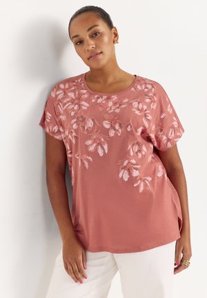 Kvinde iført en pink kortærmet top med hvide blomsterprint kombineret med hvide bukser, stående op ad en hvid væg.