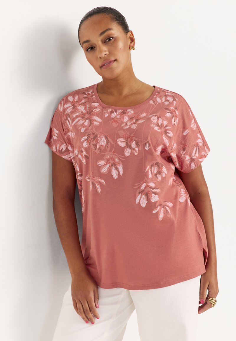 Kvinde iført en pink kortærmet top med hvide blomsterprint kombineret med hvide bukser, stående op ad en hvid væg.