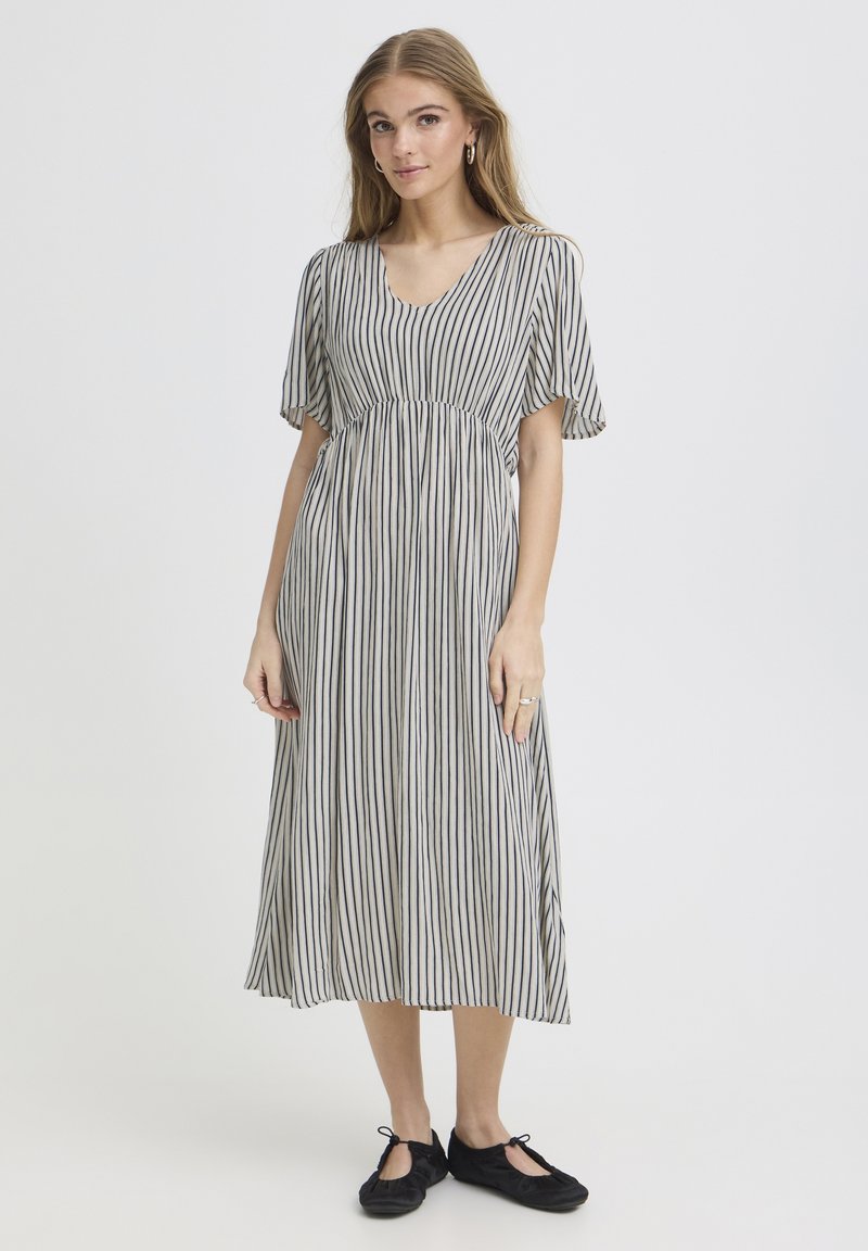 Jeune femme debout, portant une robe midi blanche à rayures noires verticales et manches volantées, associée à des ballerines noires.