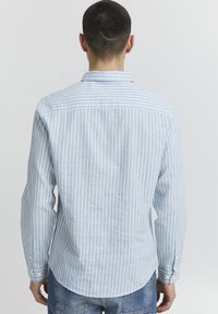 Lys blå stripete skjorte laget av stoff, med lange ermer, krage med knapper og avrundet nederkant, som viser en subtil tekstur.
