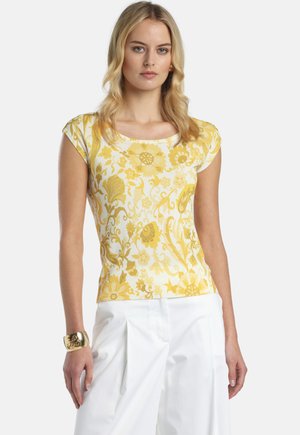 Femeie blondă purtând un top galben cu mâneci scurte, decorat cu flori, pantaloni albi plisati și o brățară aurie pe încheietura mâinii stângi.