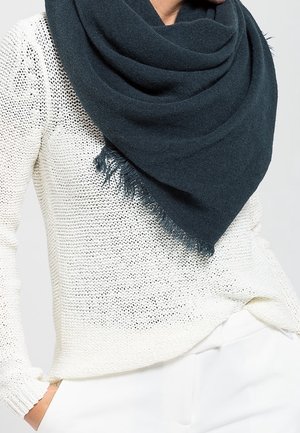 Personne portant une écharpe à franges bleu marine foncé sur un pull en maille blanc et un pantalon blanc, la main glissée dans la poche.
