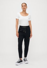 Haut blanc à encolure dégagée, pantalon noir à taille élastique avec cordon doré, et baskets noires à plateforme avec semelles blanches. Design simple et moderne.