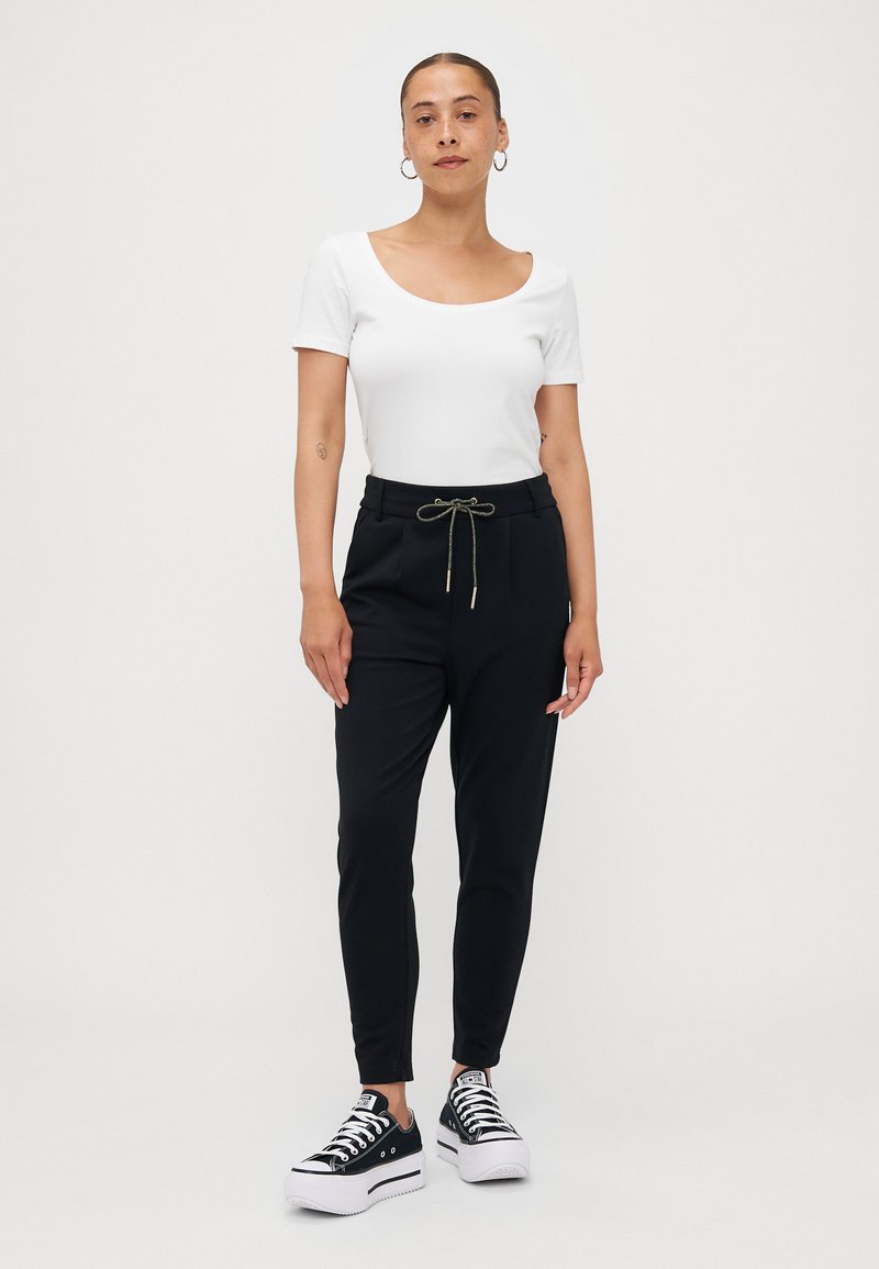 Weißes Top mit Rundhalsausschnitt, schwarze Hose mit elastischem Bund und goldener Kordel, sowie schwarze Plateausneaker mit weißen Sohlen. Einfaches und modernes Design.