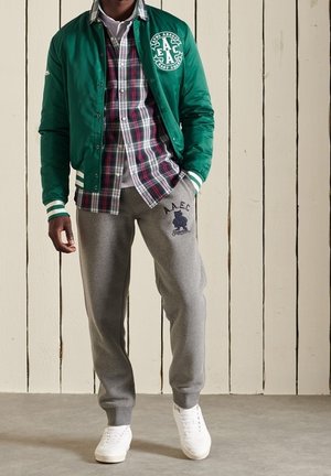 Zielona kurtka bomberka z białymi paskowanymi mankietami, nałożona na kraciastą koszulę. Szare spodnie dresowe z granatowym logo, zestawione z białymi tenisówkami.