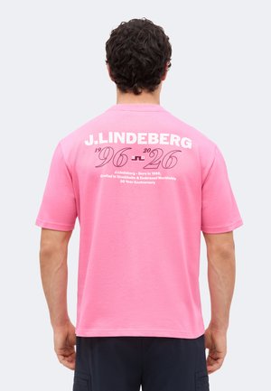 Hombre con una camiseta rosa con texto blanco "J.LINDEBERG 1996-2026 30 Year Anniversary" en la espalda, de pie de espaldas.
