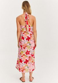 Žena v ružových kvetinových halenkových šatách s červenými, oranžovými a zelenými kvetmi, stojaca bosá chrbtom k jednofarebnému pozadiu.