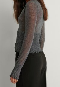 Femme portant un haut gris moulant transparent à manches longues avec une taille et des poignets texturés, associé à un pantalon noir taille haute.