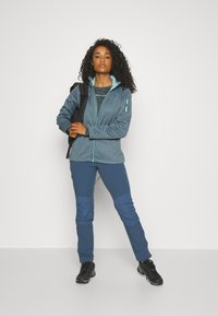 Woman in blue outdoor jacket and pants stands with black backpack slung over one shoulder on plain white background.