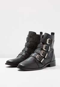 Botas de tobillo de cuero negro con un acabado texturizado, que presentan tres hebillas plateadas y una cremallera lateral. Tacón bajo y resistente para mayor estabilidad.