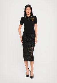 Woman wearing a black polo shirt with gold trim and emblem, paired with a black floral lace midi skirt and black pointed heels, standing.