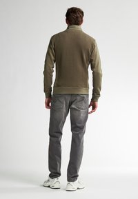 Khaki ritsjack met textuur op de rug, geribbelde manchet en bijpassende grijze jeans. Model draagt witte sportschoenen. Effen achtergrond.