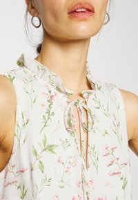 Blusa floreale in tessuto leggero e trasparente, con colletto arricciato, chiusura a lacci e delicati motivi botanici in rosa e verde.
