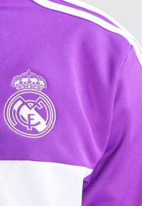Lila sporttröja med vita ränder, texturerad tyg och en broderad Real Madrid-logotyp med en krona ovanför.