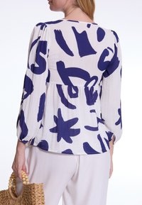 Witte top met abstracte marineblauwe patronen, lange mouwen, gathered schouderdetails en peplumstijl, gecombineerd met lichte broek en een geweven tas.