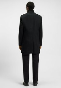 Cappotto di lana nero con colletto alto, vestibilità sartoriale e apertura centrale, abbinato a pantaloni neri coordinati e scarpe nere eleganti.