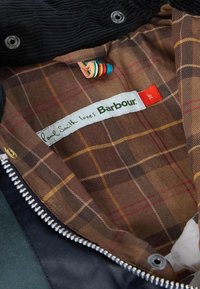 Barna kockás bélés sárga és piros részletekkel, fehér címkével, rajta a felirat: "Paul Smith szereti a Barbour-t." Ezüst cipzár és sötét szövet.