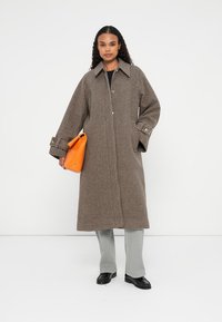 Brun houndstooth lang frakke med knaplukning, stor krave og brede ærmer, parret med grå bukser og en orange håndtaske.