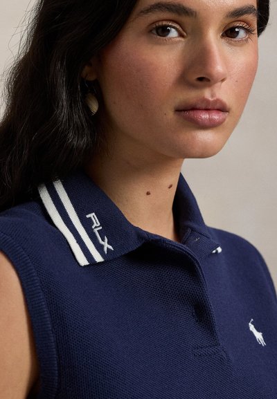 Μπλε σκούρο μπλούζα πόλο χωρίς μανίκια με υφασμένο υλικό, με λευκή ριγωτή γιακά και λογότυπο RLX, σε συνδυασμό με μικρά σκουλαρίκια.