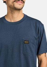 T-shirt blu navy a maniche corte in cotone misto, con taschino sul petto decorato da un'etichetta gialla con il logo RVCA. Texture liscia e design classico a girocollo.