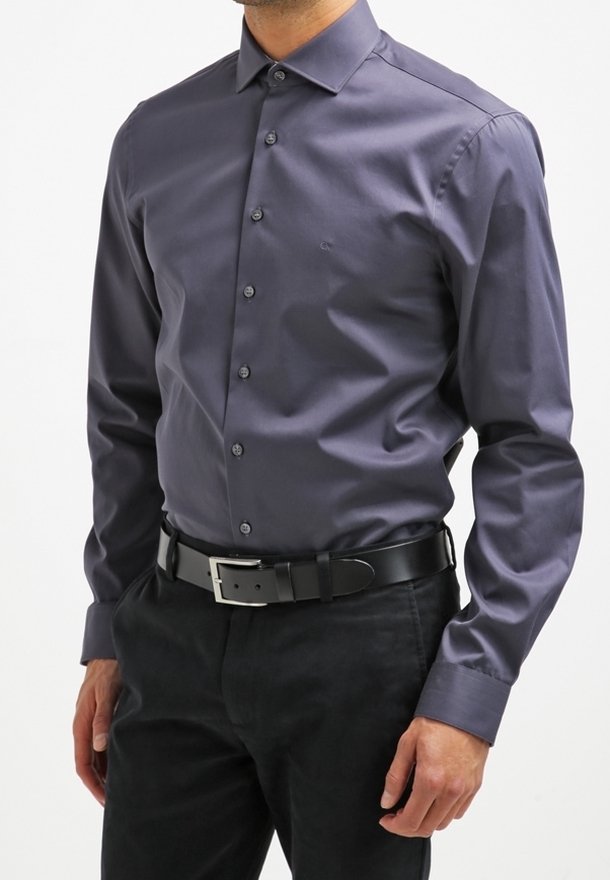 Camicia a maniche lunghe grigia scura con chiusura a bottoni, colletto e texture liscia, abbinata a una cintura nera e pantaloni scuri.