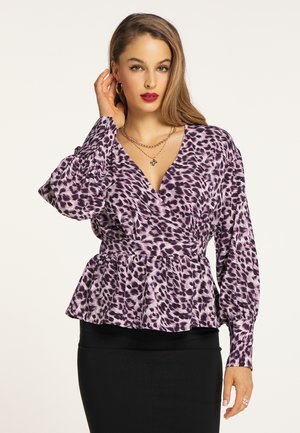 Woman with long hair wearing a purple leopard print blouse with puffed sleeves and a black skirt, accessorized with gold jewelry.