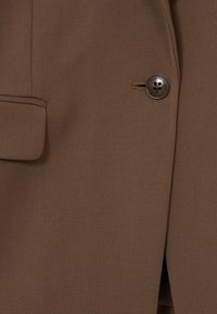 Tissu de blazer marron sur mesure avec un seul bouton foncé fermé, montrant un détail de poche à rabat sur le côté gauche.