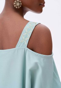 Jasnoturkusowy top z odkrytymi ramionami o gładkiej fakturze i ozdobnym złotym haftem na ramiączku, charakteryzujący się luźnym krojem i kobiecą sylwetką.
