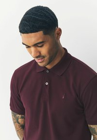 Burgundy lyhythihainen polo-paita, joka on valmistettu hengittävästä kankaasta. Paitassa on kaksinappinen nauha ja brodeerattu logo rinnassa.