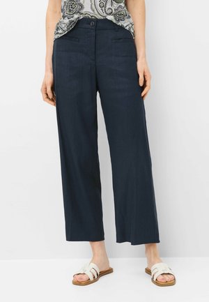 Vrouw draagt marineblauwe brede cropped broek, witte blouse met patroon ingestopt en witte slippers, staand tegen een effen achtergrond.