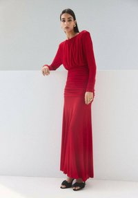 Rochie roșie cu mâneci lungi, corset adunat și fustă fluidă. Material neted cu o strălucire subtilă, asortată cu pantofi negri slip-on.