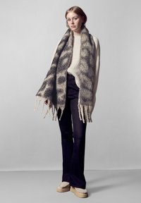 Uldscarf med et grå og beige mønster, frynset kanter, båret over en lys sweater, parret med mørke jeans og beige slip-on sko.