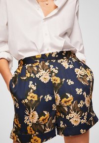 Valkoinen napillinen paita, joka on pujotettu sinisiin kukkakuosisisiin shortseihin, joissa on käännetyt hihansuut. Henkilö seisoo käsivarret taskuissaan.