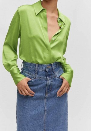 Femme portant un chemisier en satin vert citron rentré dans une jupe en denim bleu taille haute, les mains dans les poches de la jupe.