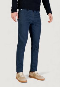 Pantaloni chinos blu navy a taglio slim, realizzati in misto cotone, con tasche frontali e chiusura con zip. Abbinati a sneakers beige.