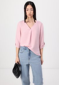 Lys rosa bluse med V-hals, trekvartlange ermer og teksturert finish, sammen med lyseblå høytlivsjeans og en svart veske.