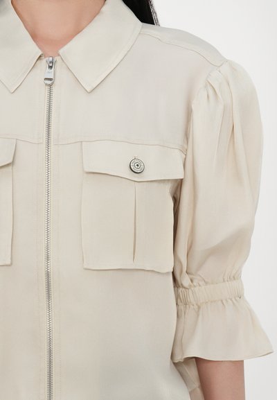 Beige tõmblukkpusa, millel on krae, puhv-sleeve'id ja kaks rinnataskut. Omab hõbedast riistvara ja pehmet, kerget kangast struktuuri.