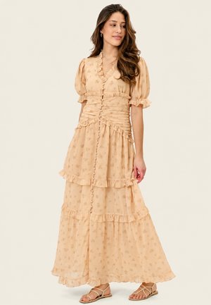 Femme aux longs cheveux ondulés, portant une robe maxi beige à fleurs avec des manches ballons et des détails à volants, debout en sandales devant un fond uni.