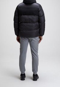 Doudoune noire avec des coutures horizontales et une capuche, associée à un pantalon gris clair avec des poches cargo et des chaussures de sport noires.