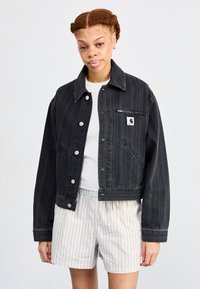 Jacket in denim nero con strisce verticali, chiusura a bottoni e tasca sul petto con etichetta logo. Indossato sopra una camicia bianca e pantaloni corti a righe.